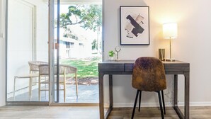 Een moderne kamer met een glazen deur naar een balkon, een bureau met stoel, een lijst met abstracte kunst, en een bureaulamp.