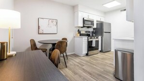 Een moderne keuken met witte kasten, een roestvrijstalen koelkast, een magnetron en een oven. Links staat een eettafel met vier stoelen en een lamp.
