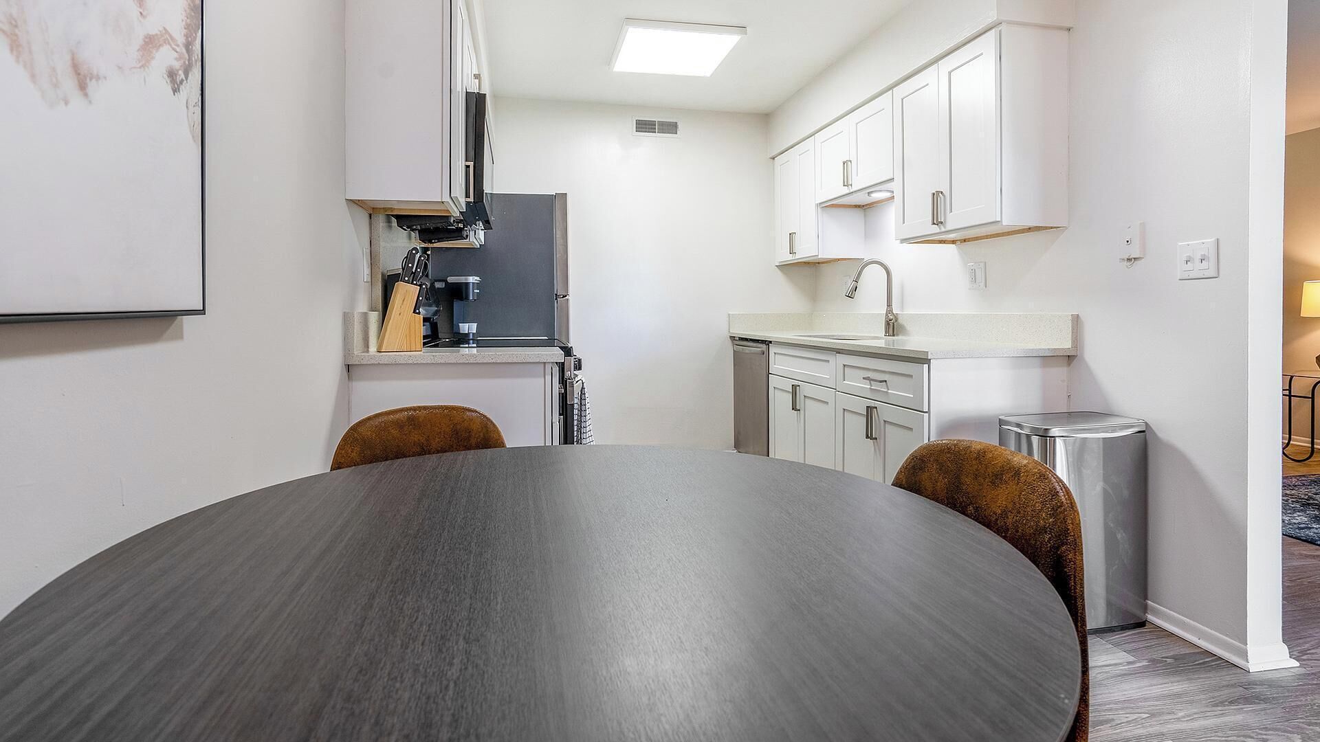 Cozinha moderna com armários brancos, forno embutido e pia. Há uma mesa de jantar com duas cadeiras em primeiro plano.