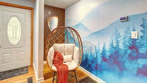 Quarto com piso de madeira, porta branca com painel de vidro decorativo, cadeira de vime com almofada e um mural na parede retratando uma floresta.