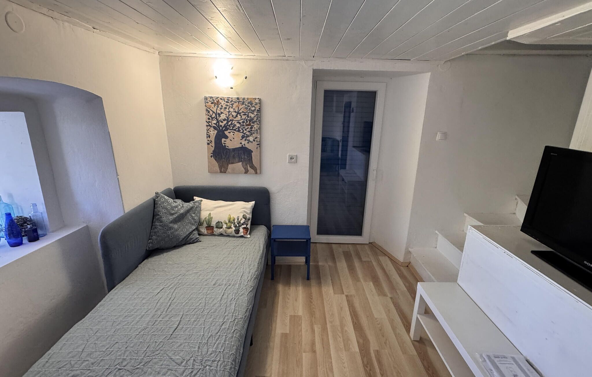 Une petite pièce bien éclairée, avec un lit, une télévision, une petite table et une porte donnant sur un balcon.
