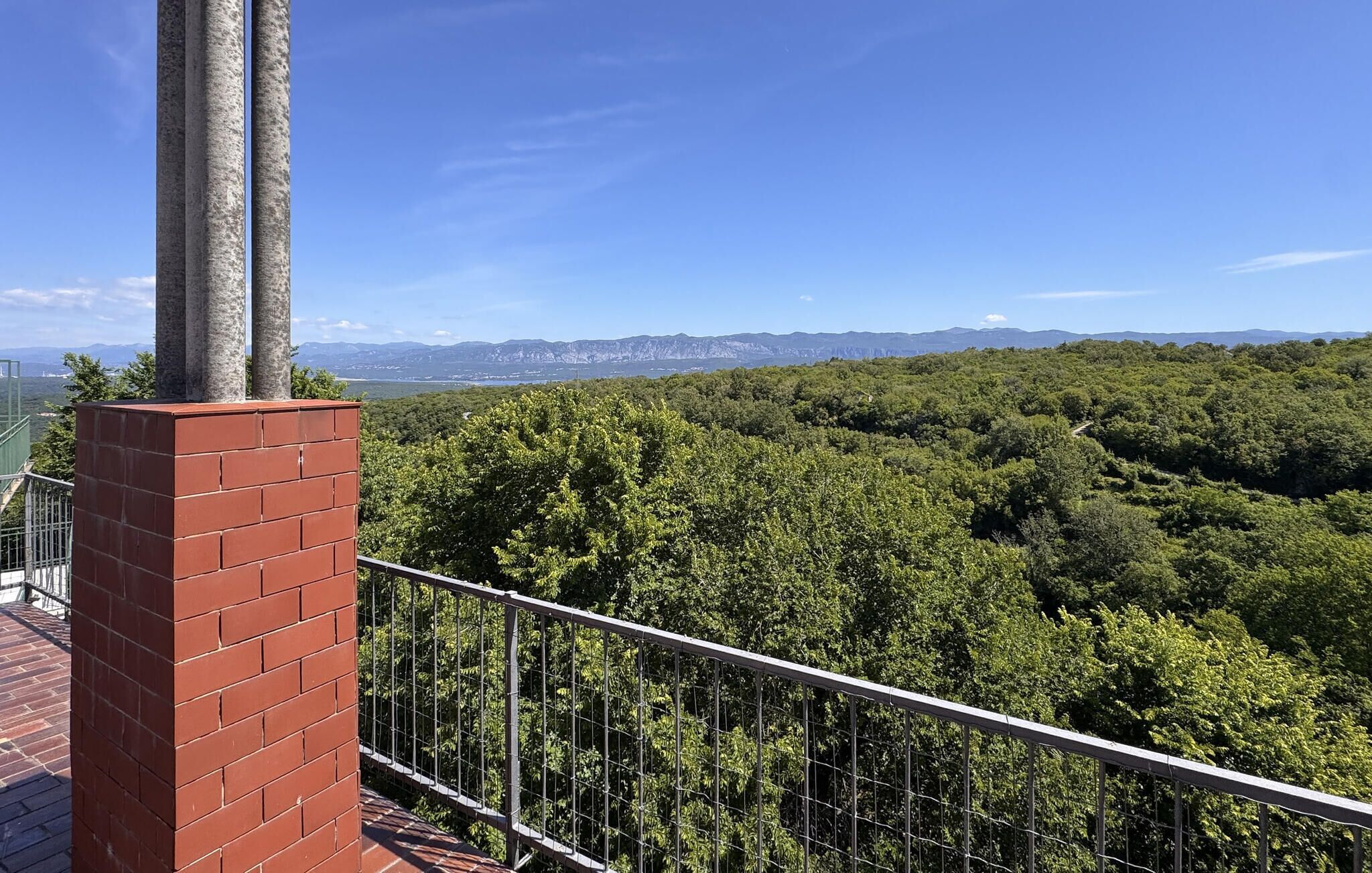 A balcony with a brick wall and metal railing overlooking a dense forest and distant mountains.