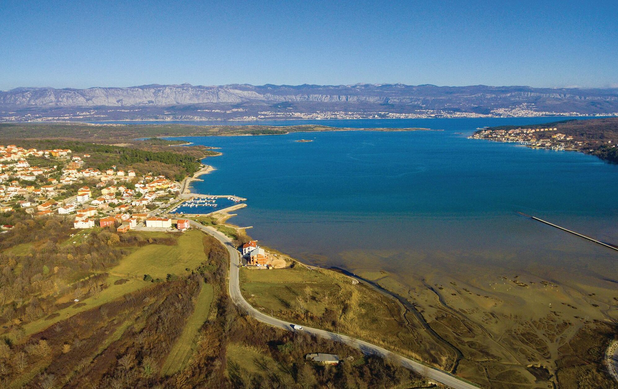Vue aérienne d’une ville côtière dotée d’un port de plaisance, entourée de montagnes et bordée par une vaste étendue d’eau.
