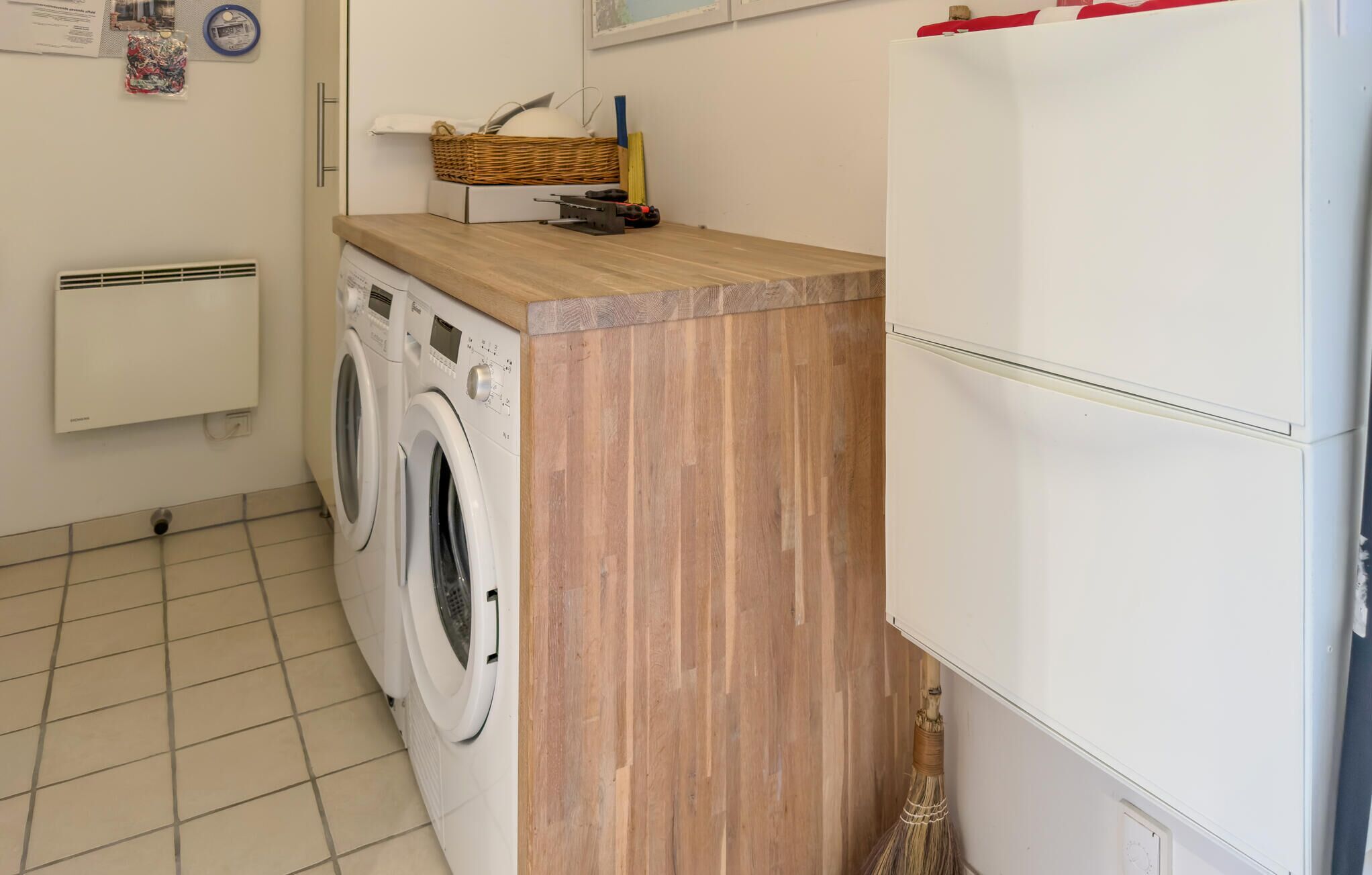 A laundry area with two washing machines, a wooden counter, a basket, and a broom.