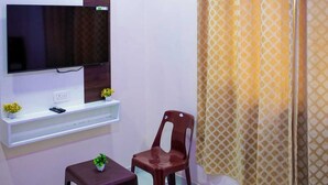 A room with a flat-screen TV mounted on the wall, a small table with a potted plant, a wooden chair, and a patterned curtain. - Hotel Saraswati Stay Dham (Vrindavan)
