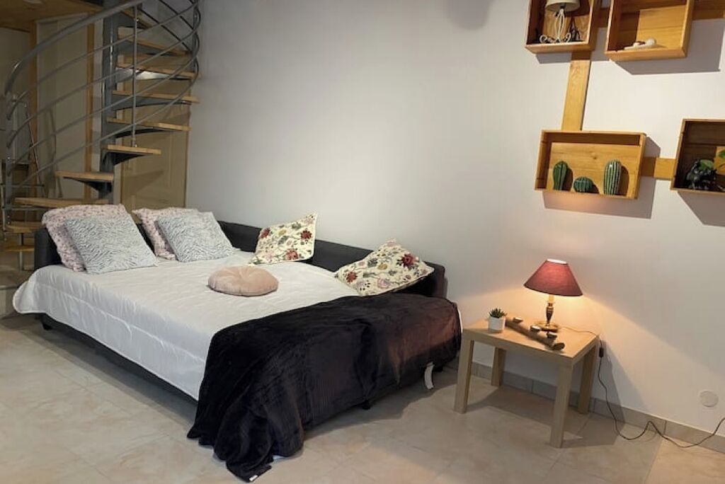 A bedroom with a bed, a nightstand, a lamp, a wall-mounted shelf with decorative items, and a spiral staircase.