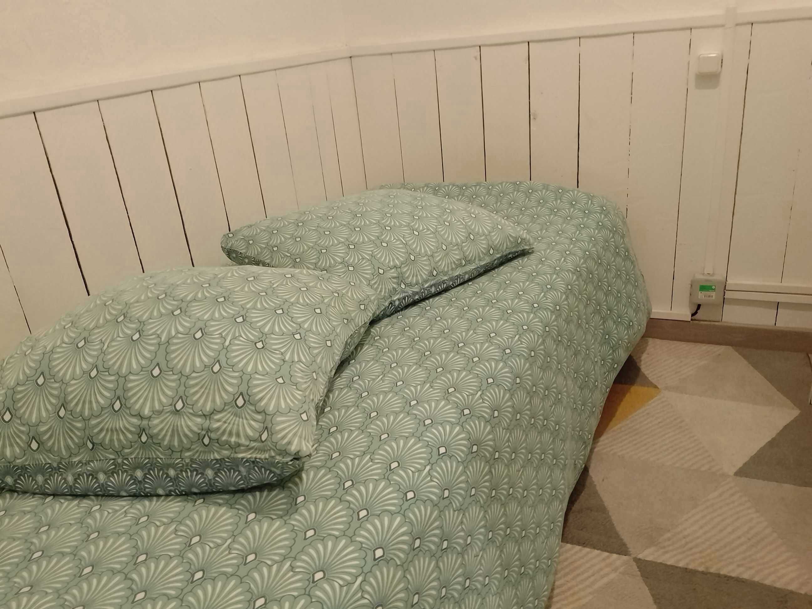 Ein Bett mit einem gemusterten Überwurf und Kissen, eine weiße Wand mit vertikaler Paneelung und ein Teppich mit geometrischem Muster.