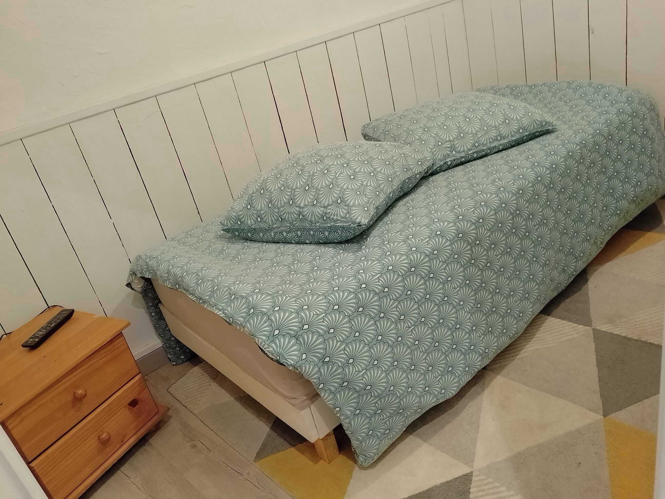 Una cama individual con colcha y almohada estampadas, una mesita de noche de madera con control remoto y un tapete de diseño geométrico sobre piso de baldosas.