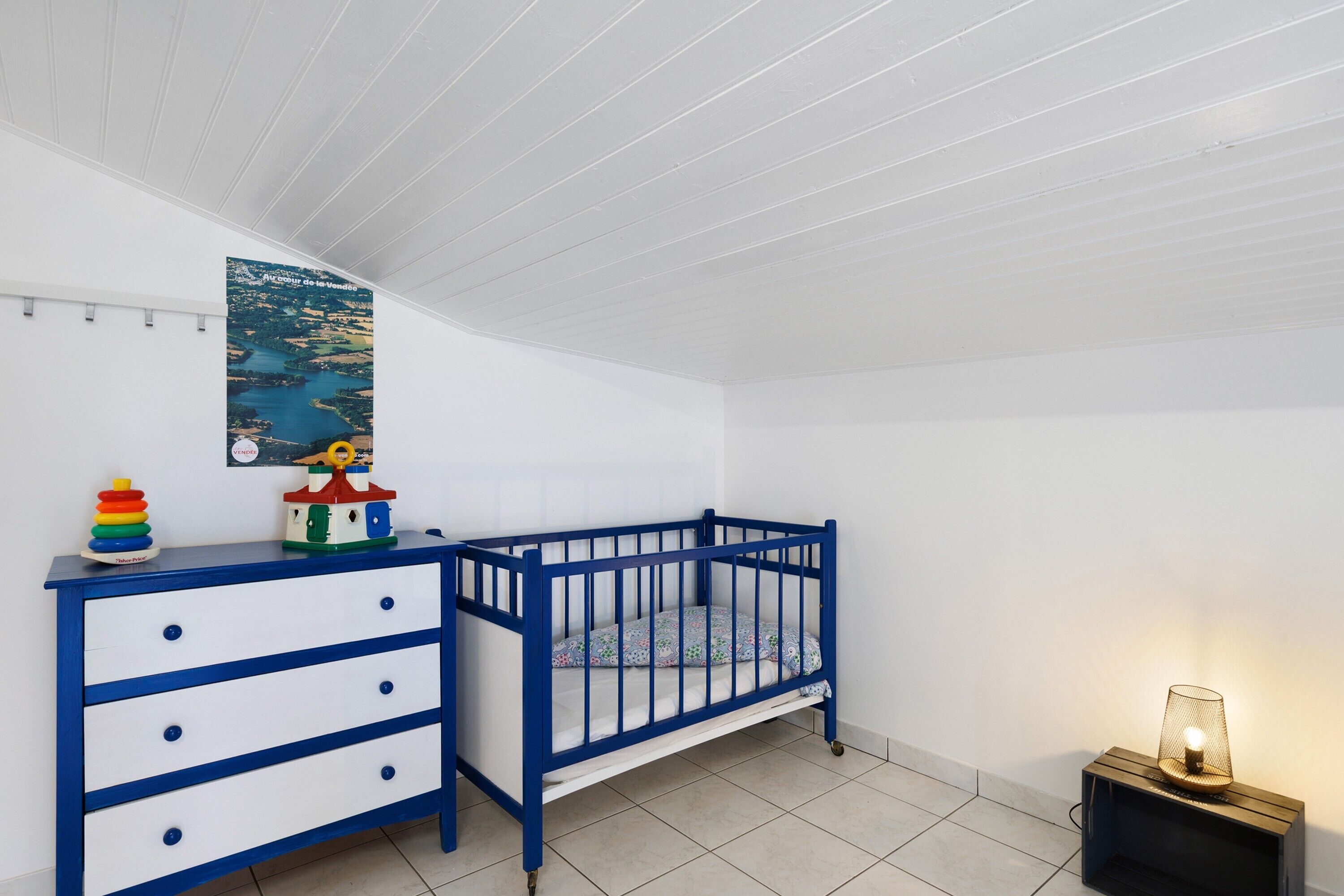 Une chambre d’enfant avec un berceau, une commode, une affiche et une lampe.