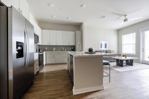 Eine moderne Küche mit einer zentralen Kochinsel, Edelstahlgeräten und einem Essbereich mit grauem Sofa und Couchtisch.