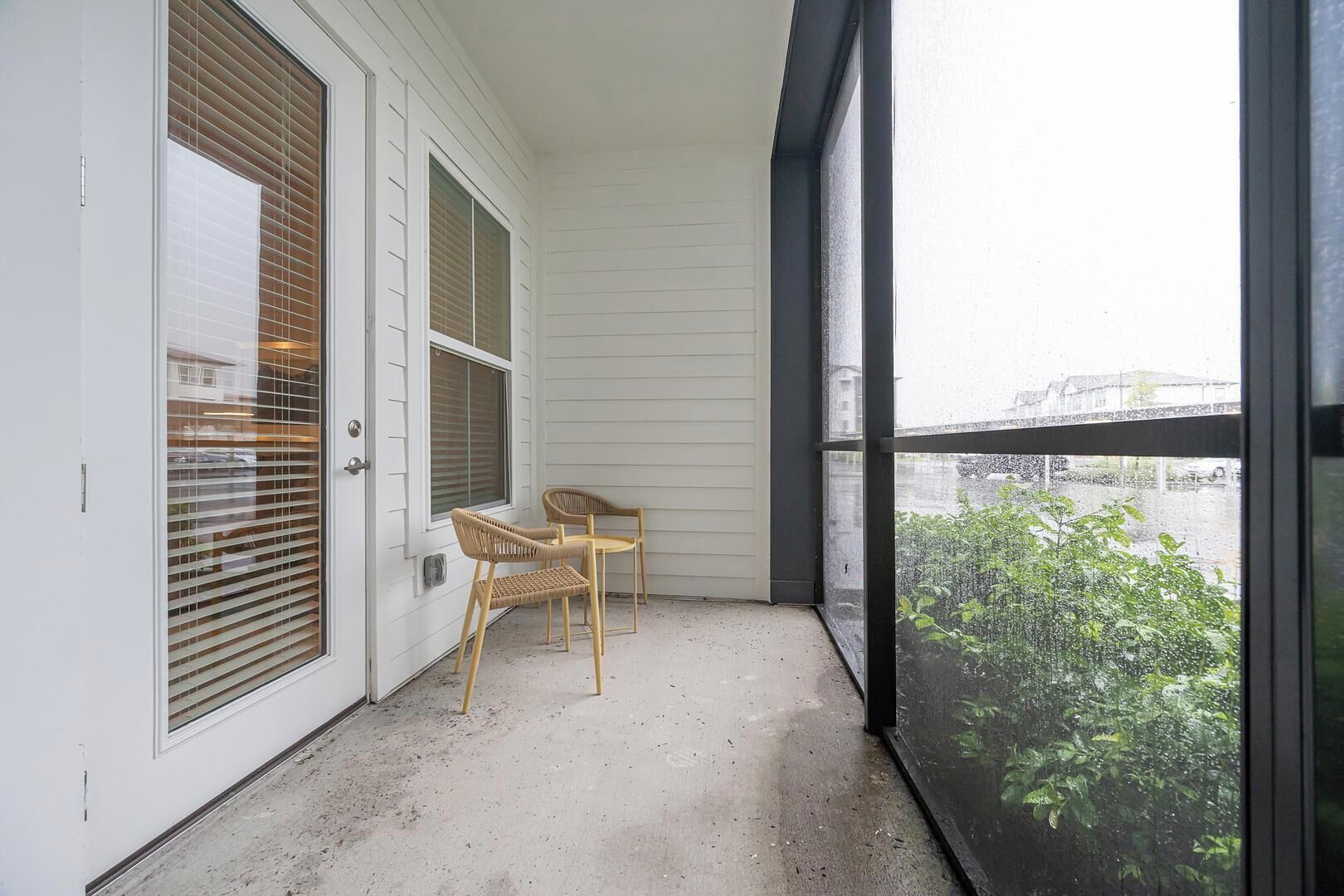 Een balkon met twee rieten stoelen, een witte deur met zonwering, en uitzicht op groen door een groot raam.