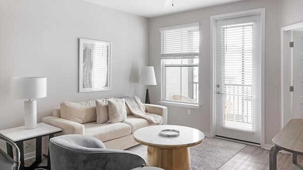 A modern living room with a beige sofa, a round coffee table, and a grey armchair. There are two windows with blinds, a lamp, and a framed picture on the wall. - Landing | Stylish 1BD, Clubhouse, Gym (Wimauma)