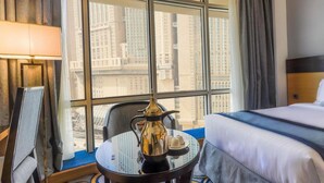 A hotel room with a bed, a table with a coffee pot, a chair, and a view of tall buildings through large windows. - Prestige Al Mashaer Hotel (Makkah)