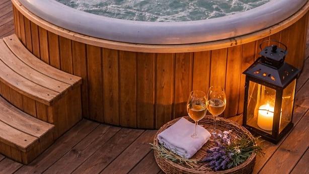 A hot tub surrounded by wooden deck seating, with two glasses of wine and a candle on a tray. - Les insolites de la bastide (La Bastide)