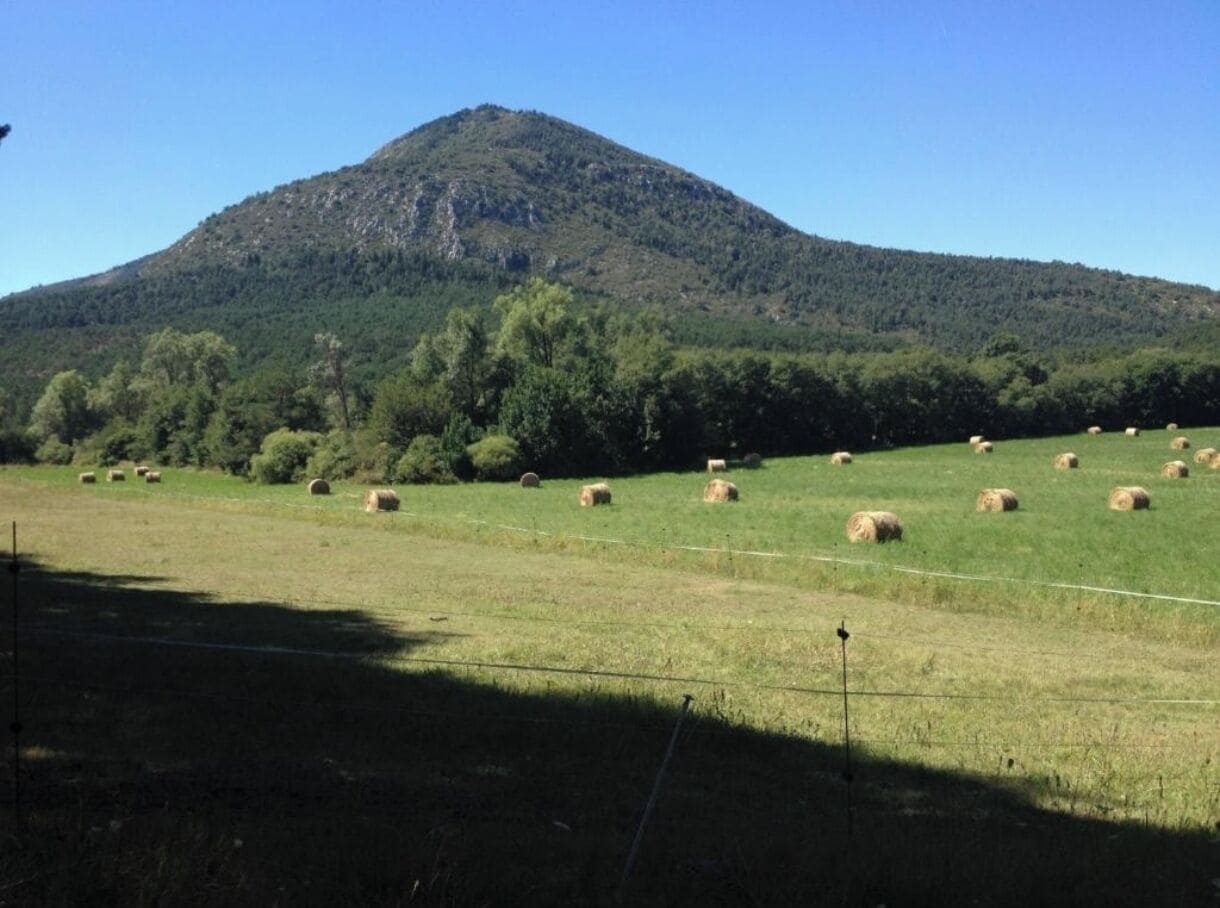 Un paisaje rural con una montaña al fondo, pacas de heno dispersas en un campo y una cerca que atraviesa la escena.