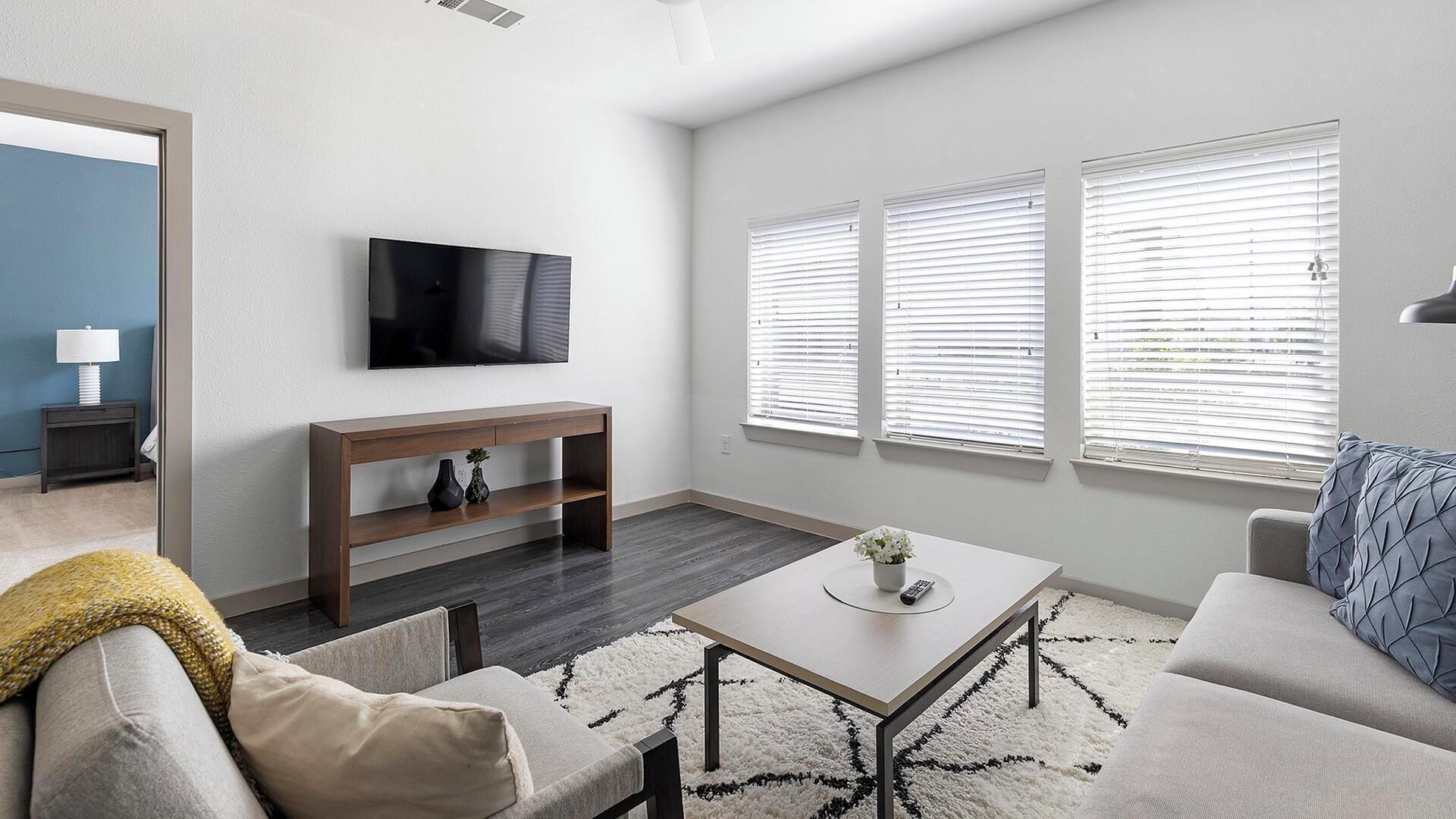 A modern living room with a flat-screen TV, a wooden TV stand, a sofa, a coffee table, and large windows with blinds.