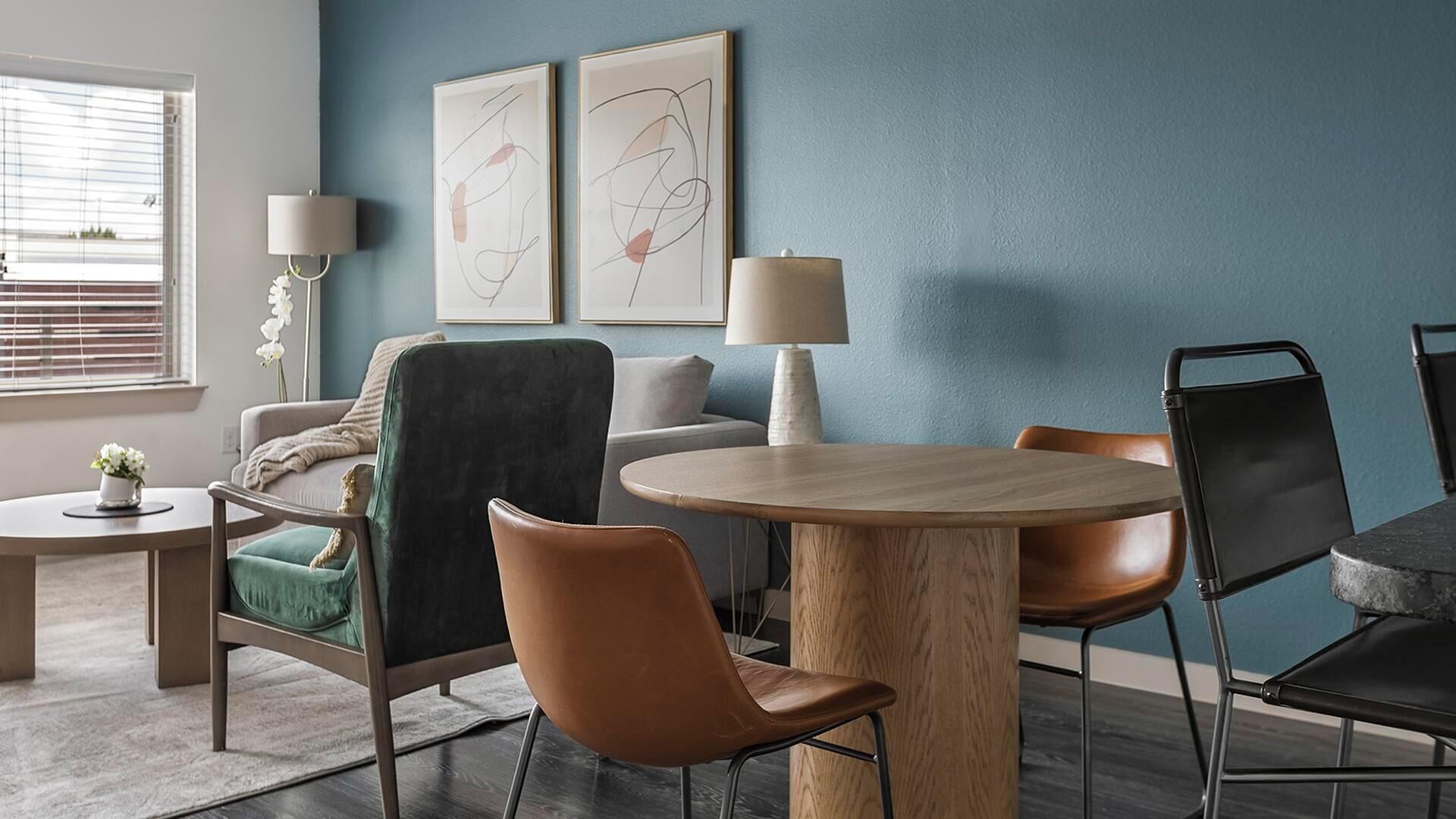 Een moderne woonkamer met een ronde houten tafel, twee stoelen, een bank en twee abstracte schilderijen aan de muur.