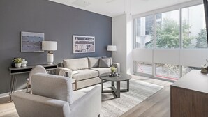 Ein modernes Wohnzimmer mit einem grauen Sofa, einem runden Couchtisch und zwei Stehlampen. An der Wand befinden sich zwei gerahmte Kunstwerke und ein Fenster mit Blick ins Freie.