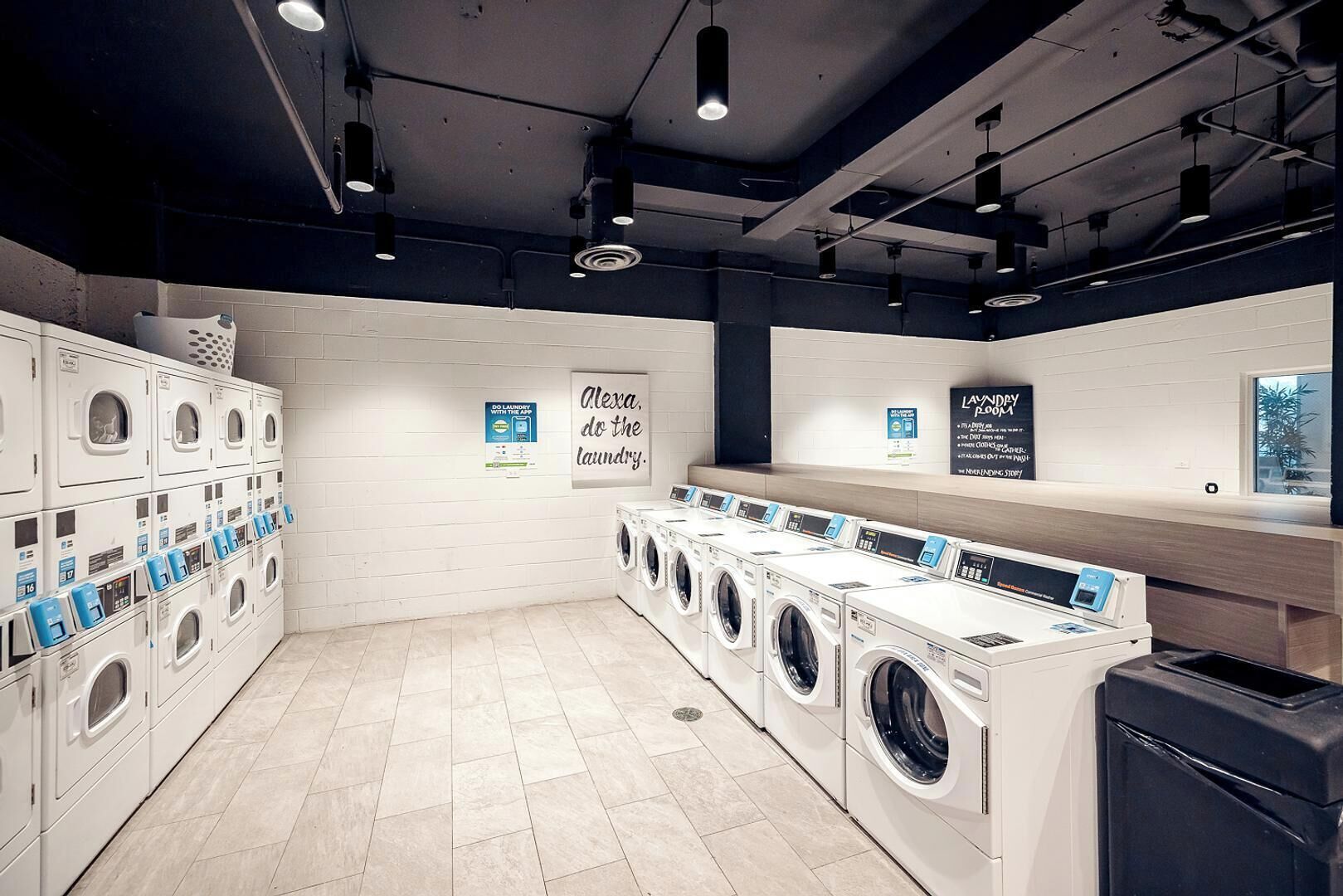 Une laverie moderne équipée de plusieurs machines à laver et de plusieurs séchoirs, d’un panneau fixé au mur et d’une poubelle.