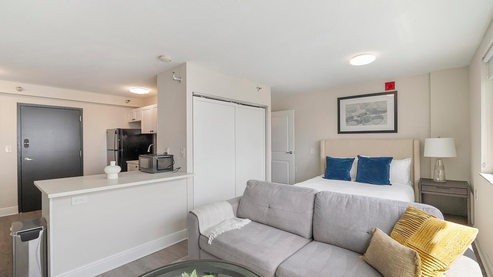 Ett kompakt hotellrum med ett litet pentry, en grå soffa, en säng med blå och beige kuddar samt ett litet nattduksbord med en lampa.