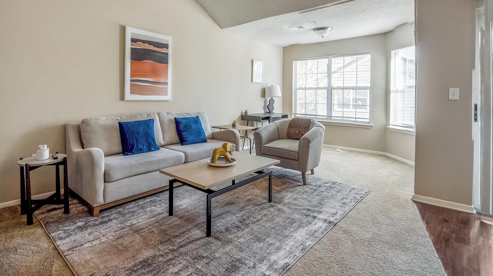 A living room with a sofa, armchair, coffee table, and a rug. There is a painting on the wall and a window with blinds.