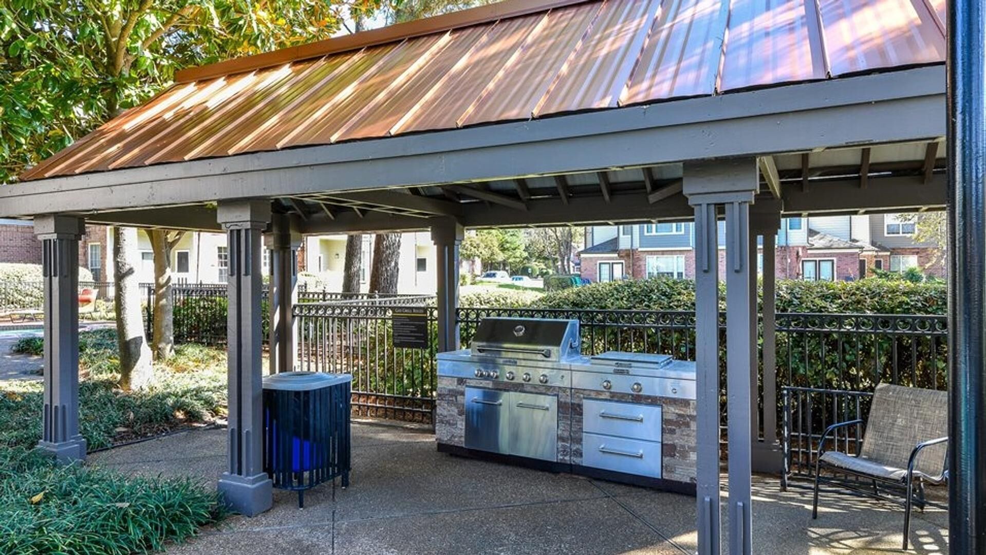 An outdoor patio with a grill and seating area, surrounded by a metal fence and trees.
