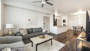 A modern living room with a grey sofa, a coffee table, and a TV. There is a kitchen area with a dining table and chairs. - Landing | Stunning 1BD, Gym (Glendale)
