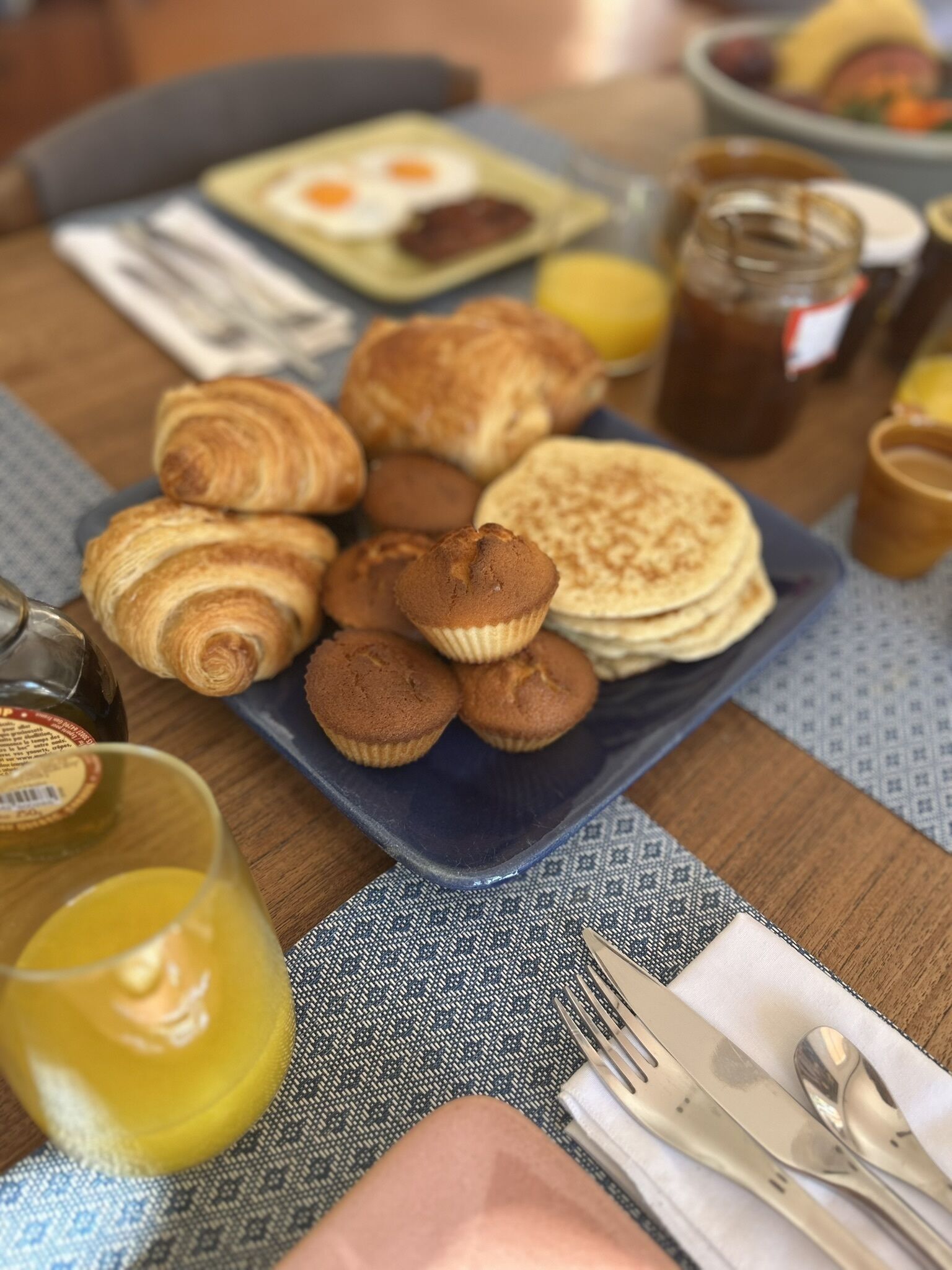 Ein Frühstückstisch mit Croissants, Muffins, Pfannkuchen, einem Glas Orangensaft und einer Marmeladenglas.