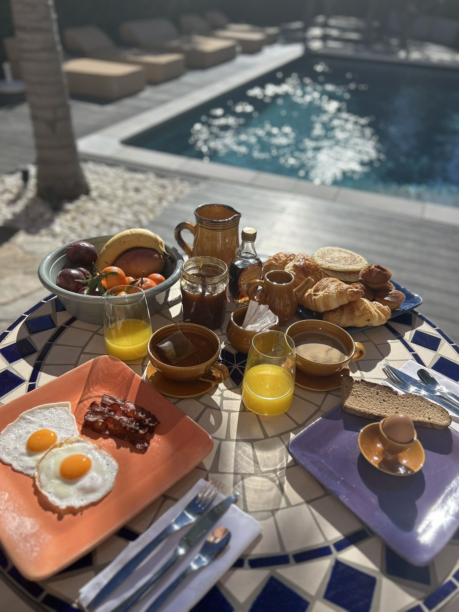 Desayuno con huevos, tocino, tostadas, pasteles y bebidas sobre una mesa con azulejos cerca de una piscina.
