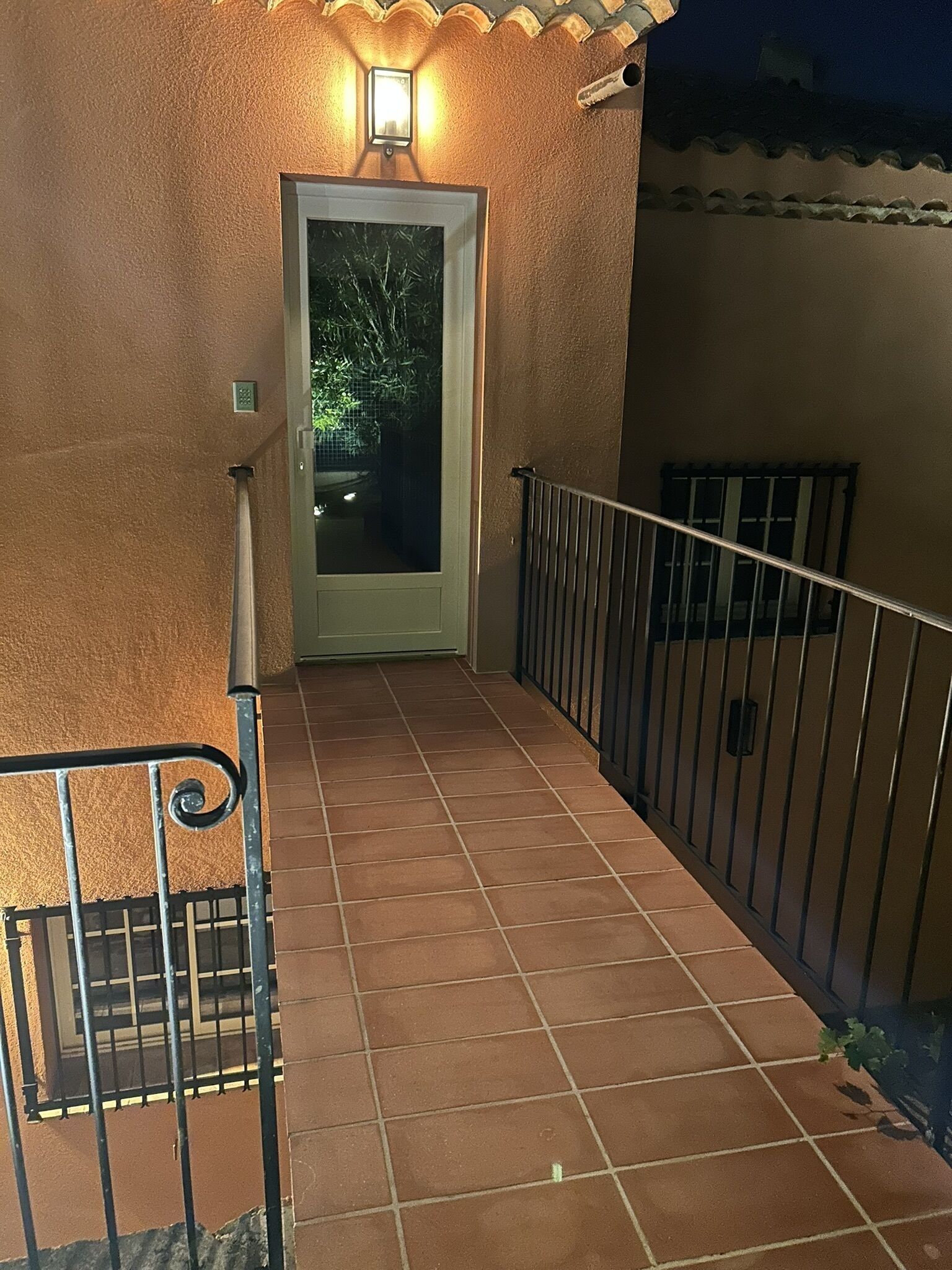 Un pasillo exterior con baldosas, barandilla de metal, una puerta con luz encima y una pared con cornisa decorativa.