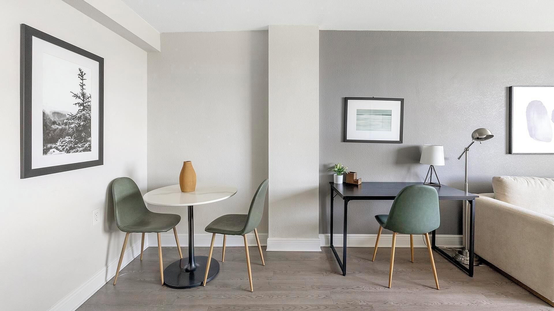 Un salon moderne avec une table à manger ronde, deux chaises, un canapé et un bureau avec une lampe. Des tableaux encadrés ornent les murs et un vase est posé sur la table.