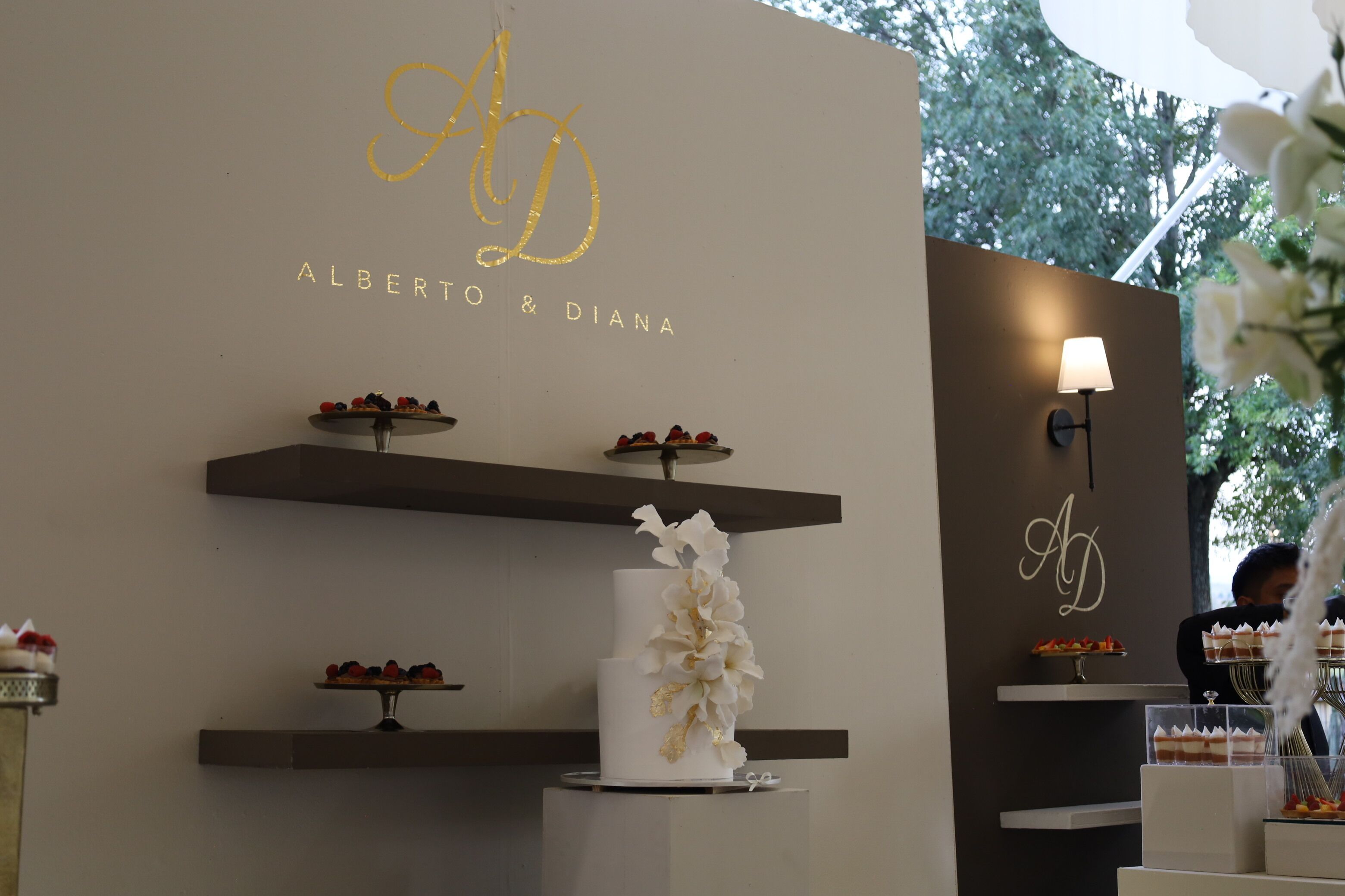 A wedding cake with floral decorations on a pedestal, surrounded by dessert trays on shelves.