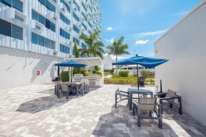Une terrasse extérieure avec des tables et des chaises, un barbecue et un bâtiment blanc en arrière-plan.