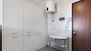Una kitchenette con un armario blanco, una mesita con una silla, un calentador de agua y una puerta con cerradura.