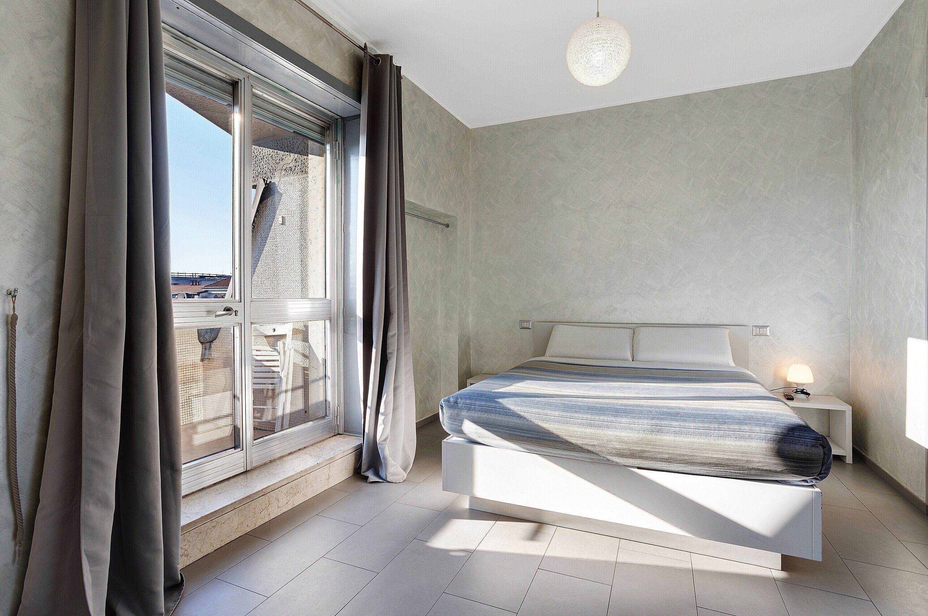 A modern bedroom with a large bed, a window with curtains, a bedside table with a lamp, and a view of the outside.