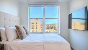 A bedroom with a large window, a bed with white bedding, and a mounted TV on the wall.