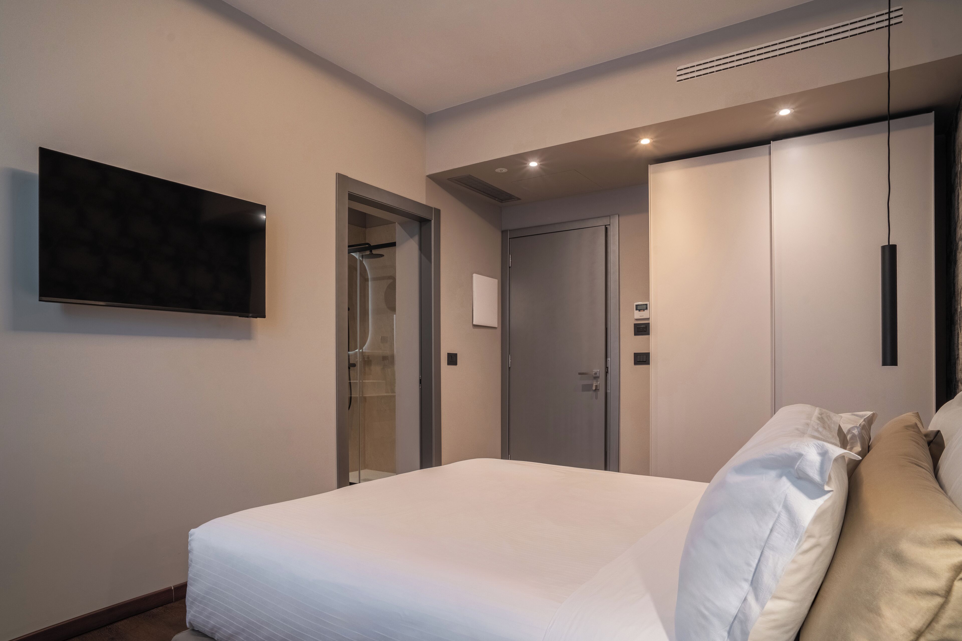 A modern hotel room with a large bed, a flat-screen TV mounted on the wall, a wardrobe, and a walk-in closet.