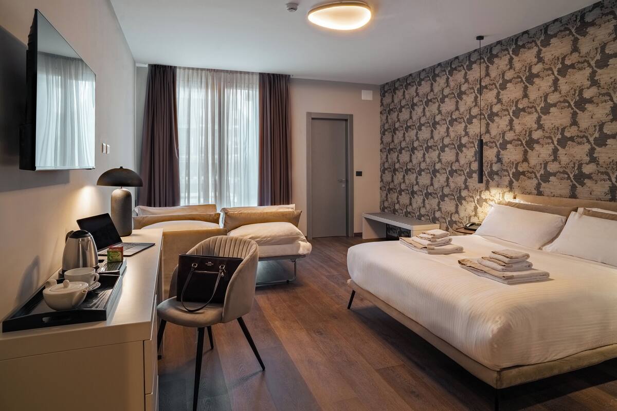 Ein Hotelzimmer mit Bett, Schreibtisch mit Laptop, Stuhl, Nachttisch mit Lampe und Kleiderschrank.