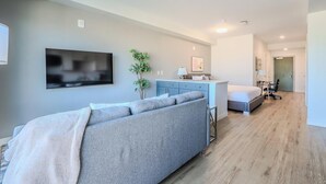 A modern living room with a grey sofa, a flat-screen TV, a small kitchen area, and a bedroom with a bed and desk.