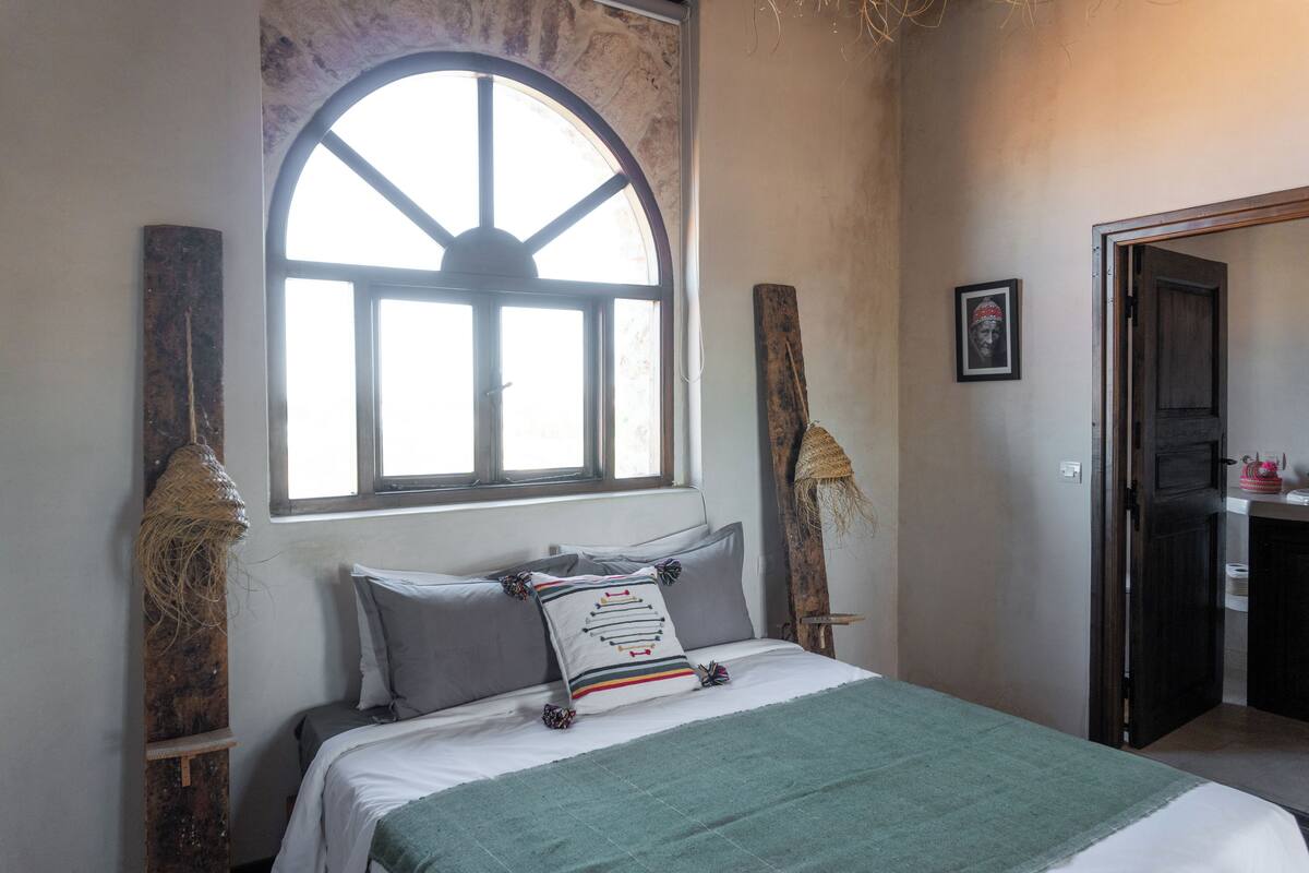 Une chambre avec une grande fenêtre en arc de cercle, un lit avec une literie grise et un plaid vert.