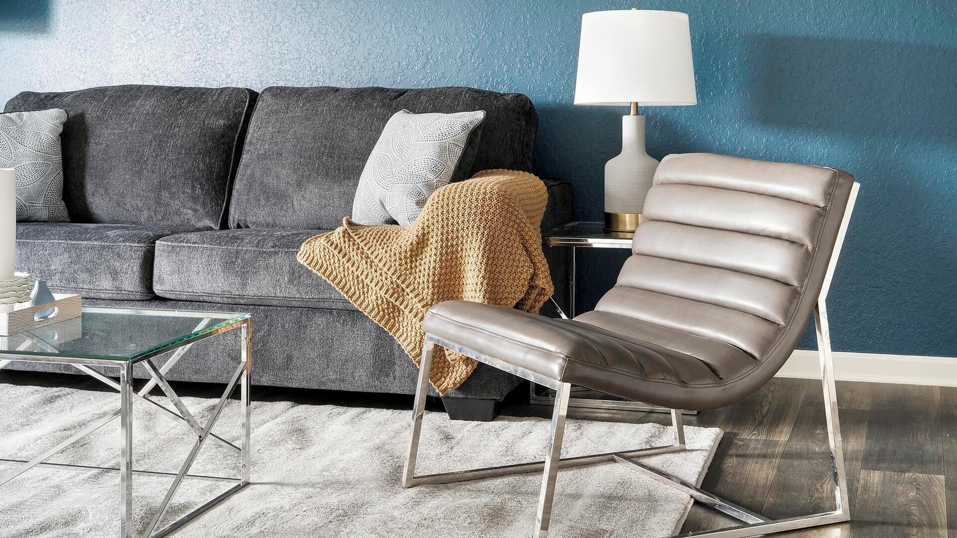 Ein modernes Wohnzimmer mit grauem Sofa, Ledersessel, Glastisch und einer Lampe.
