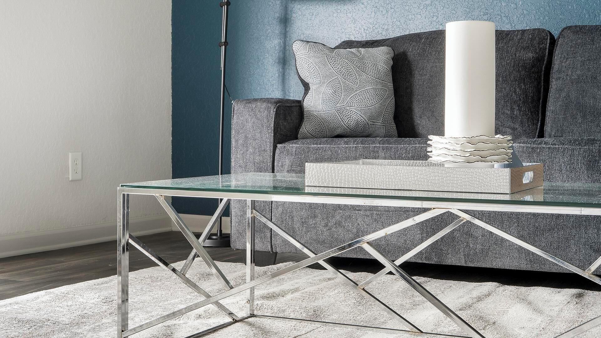 A modern living room with a glass coffee table, a grey sofa with patterned cushions, and a floor lamp.