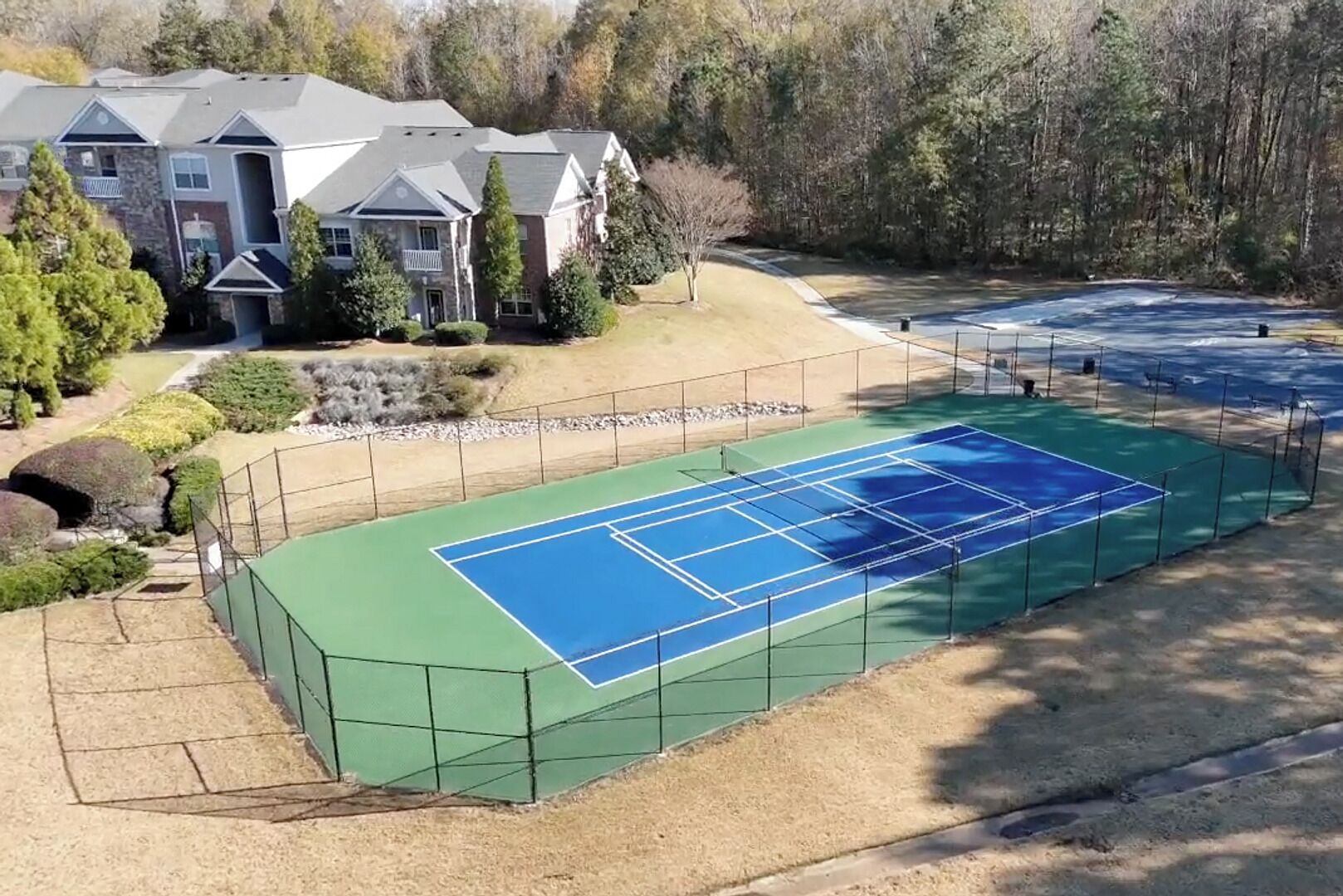 Una cancha de tenis con superficie verde y azul, rodeada por una valla, con una casa y árboles al fondo.