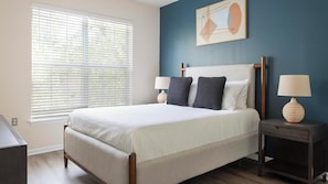 A bedroom with a bed, two bedside tables with lamps, a window with blinds, and a wall art piece.