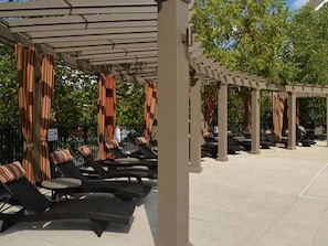 A covered outdoor seating area with lounge chairs and tables, a pergola structure, and a paved floor.