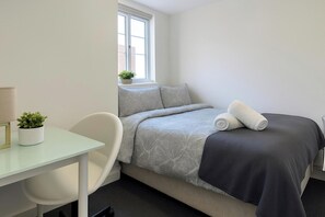 A small, neatly arranged bedroom with a bed, a desk, and a chair. There is a window with a view of buildings outside. - 7 Bed House - Sleeps 14 - Parking (Bournemouth)