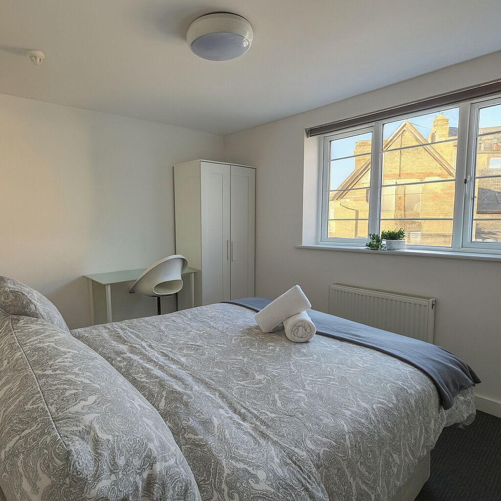 A bedroom with a bed, a desk, a chair, a wardrobe, and a window with a view of buildings.