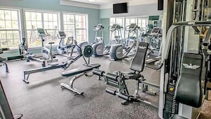 Een sportschool met diverse fitnessapparaten, grote ramen en plafondventilatoren.