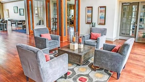 Een ruime lobby met een hoge plafond, houten balken en een centrale salontafel omringd door grijze fauteuils.