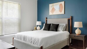 Een slaapkamer met een bed, twee nachtkastjes, een raam met jaloezieën en een aan de muur hangend abstract schilderij.